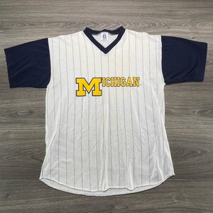 May include: A short-sleeve V-neck shirt featuring a white base with thin vertical navy blue pinstripes and solid navy blue sleeves. The front displays the word MICHIGAN in yellow block letters with a large yellow M on the left side.