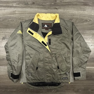 May include: A gray and yellow Nike ACG jacket with a black collar and accents. The jacket has a zipper closure, multiple pockets, and the Nike ACG logo on the sleeve. The jacket also has the text "NIKE FIT" on the bottom.