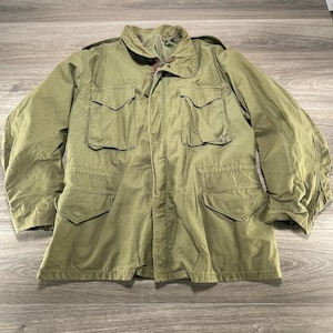 May include: An olive green field jacket featuring a high collar, a front zipper closure, and four large flap pockets on the front. The jacket includes shoulder epaulets and long sleeves, constructed with a durable fabric and a structured design.