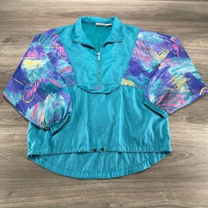 Könnte beinhalten: Eine türkisfarbene Reebok-Windjacke mit halbem Reißverschluss und einem lebendigen, mehrfarbigen abstrakten Druck auf den Ärmeln und der oberen Brust. Das Kleidungsstück verfügt über lange Ärmel mit elastischen Bündchen, einen hohen Kragen und einen verstellbaren Saum.