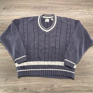 May include: A long-sleeve V-neck jumper in a muted blue-grey colour featuring a vertical cable knit pattern. The garment includes white and blue-grey striped ribbed trim along the neckline, cuffs, and hem, with a GAP brand label at the neck.