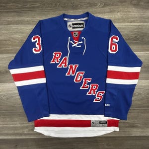 May include: A blue Reebok New York Rangers hockey jersey featuring the text RANGERS in red and white lettering across the chest, the number 36 on the sleeves, a white lace-up collar, and red and white horizontal stripes on the sleeves and hem.