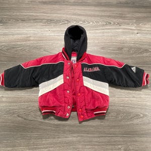 May include: A red and black hooded jacket featuring the word ALABAMA embroidered in white on the chest. The jacket has a black hood, long sleeves with red and white striped cuffs, a snap-button front closure, and white diagonal accent stripes.