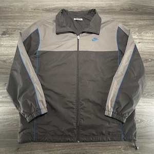 May include: A two-tone zip-up jacket featuring a dark charcoal lower body and a light gray upper chest and collar. The jacket includes long sleeves with blue piping accents, a full-length front zipper, and a small blue Nike logo on the chest.