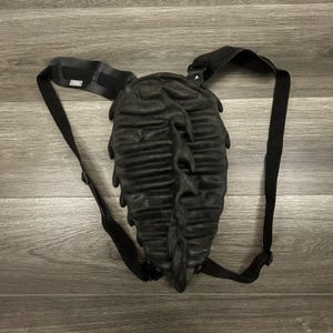 May include: A black backpack with a textured, ridged, leaf-like shell design. The bag features adjustable black nylon straps with a small rectangular tag labelled "bug" on the left shoulder strap. The central body has a distinct, organic, spine-like vertical ridge running down the middle of the shell.