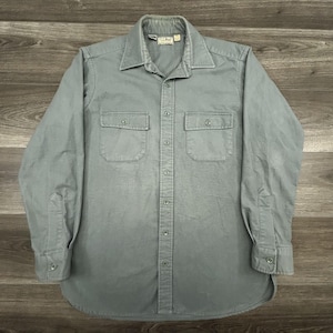 May include: A long-sleeved, button-down shirt in a muted green-gray color. The shirt features two chest pockets, a collar, and buttoned cuffs. The L.L.Bean label is visible on the collar. The shirt is made of a durable fabric.