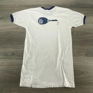May include: A white ringer t-shirt with dark blue trim on the collar and sleeve cuffs. The chest features a dark blue graphic of a racquetball racket with the text The Back Wall printed in white inside the head of the racket.