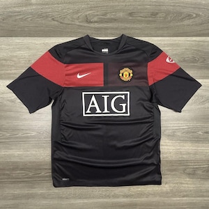 Może przedstawiać: Czarna i czerwona koszulka piłkarska z krótkim rękawem z białym logo AIG. Koszulka ma logo Nike i herb drużyny na piersi. Rękawy mają czerwony, teksturowany wzór.