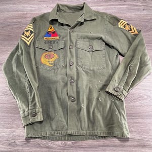 Könnte beinhalten: Ein olivgrünes, langärmeliges Hemd mit Knopfleiste und militärisch anmutenden Aufnähern, darunter ein dreieckiges Hell on Wheels-Emblem, ein 49ers-Logo und Ärmelabzeichen. Das Kleidungsstück weist sichtbare Gebrauchsspuren mit kleinen Löchern auf.