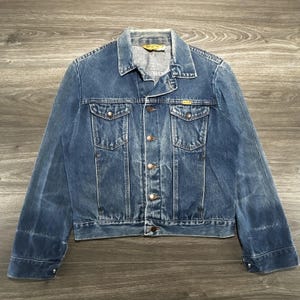 May include: A medium-wash blue denim jacket featuring a classic collar, long sleeves with buttoned cuffs, and a front button closure. The jacket includes two chest pockets with buttoned flaps and a small yellow brand tag on the left chest pocket.