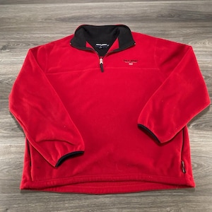 May include: A bright red fleece pullover with a black mock neck collar and a quarter-zip closure. The garment features the text Polo Sport embroidered in black on the left chest and black trim on the sleeve cuffs and collar.