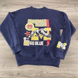 Può includere: Una felpa girocollo blu navy con una stampa grafica di un ghiottone, il testo MICHIGAN WOLVERINES e GO BLUE. Il design include una M gialla a blocchi, una stella rossa e un emblema circolare. Un piccolo ghiottone è presente sulla manica sinistra.