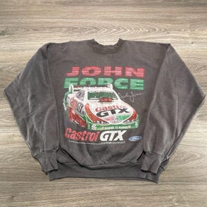 May include: A charcoal gray crewneck sweatshirt featuring a graphic print of a John Force Castrol GTX Ford Mustang race car. The design includes red and green text, a signature, and the Castrol GTX logo on the front of the long-sleeve garment.
