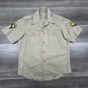 May include: A beige short-sleeved shirt with two chest pockets and button fastenings. The shirt features epaulettes and patches with a yellow emblem on each sleeve. The shirt is made of a robust fabric and has a classic, utilitarian design.
