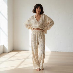 Puede incluir: Conjunto de pijama de dos piezas color crema con un delicado estampado floral. El conjunto incluye una camisa abotonada con mangas con volantes y pantalones de pernera ancha. La tela parece ligera y cómoda, adecuada para dormir o descansar.