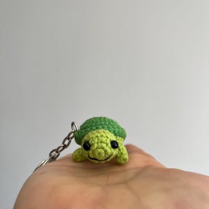 May include: A small, handmade crochet turtle keyring featuring a bright green shell and a lighter lime green head and limbs. The turtle has two small black bead eyes and a stitched black thread mouth, attached to a silver-toned metal chain.