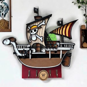 Op de afbeelding: Een houten sleutelhouder voor aan de muur in de vorm van een schip. Het schip heeft een bruine romp, witte zeilen met een doodshoofd en gekruiste botten en een rode basis met sleutelhaken. Er is ook een cirkelvormig embleem met een doodshoofd en gekruiste botten.