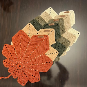 May include: A collection of handmade crochet leaf-shaped trivets in various fall colors. The trivets are in shades of orange, green, and beige. Each trivet has a unique design and is made of soft yarn. The trivets are perfect for protecting surfaces from hot dishes.