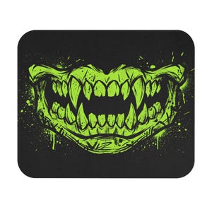 Puede incluir: Una alfombrilla de ratón rectangular con fondo negro que presenta un gráfico verde lima de una boca de monstruo estilizada con colmillos y dientes afilados. El diseño incluye efectos de salpicaduras de pintura y las letras VIZ impresas.