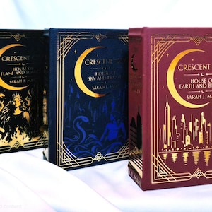 Könnte beinhalten: Drei gebundene Bücher aus der Crescent City-Reihe von Sarah J. Maas. Das schwarze Buch zeigt einen goldenen Halbmond und eine Figur mit Schwert. Das blaue Buch zeigt einen Halbmond und eine Schlange. Das weinrote Buch zeigt eine goldene Skyline.