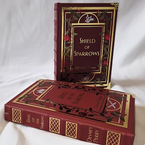 Op de afbeelding: Twee hardcover boeken met bordeauxrode covers en gouden accenten. Eén boek staat rechtop en toont "Shield of Sparrows" met bloemen- en vogelillustraties. Het andere boek, plat liggend, heeft de titel "Rites of the Starling".