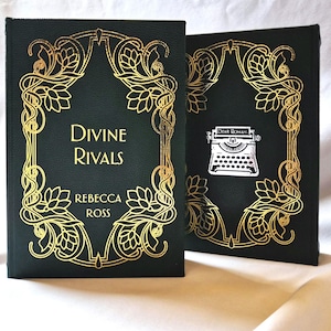 May include: Two hardback books with dark green covers and gold foil designs. The front cover of the first book reads "Divine Rivals" by Rebecca Ross. The second book has a typewriter graphic and the text "Dear Roman".