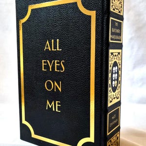 Puede incluir: Un libro de tapa dura negro con acabado texturizado y letras doradas en la portada que dicen ALL EYES ON ME. El lomo presenta paneles decorativos dorados, el t&iacute;tulo THE BUTCHERS MASQUERADE y el nombre del autor MATT DINNIMAN.