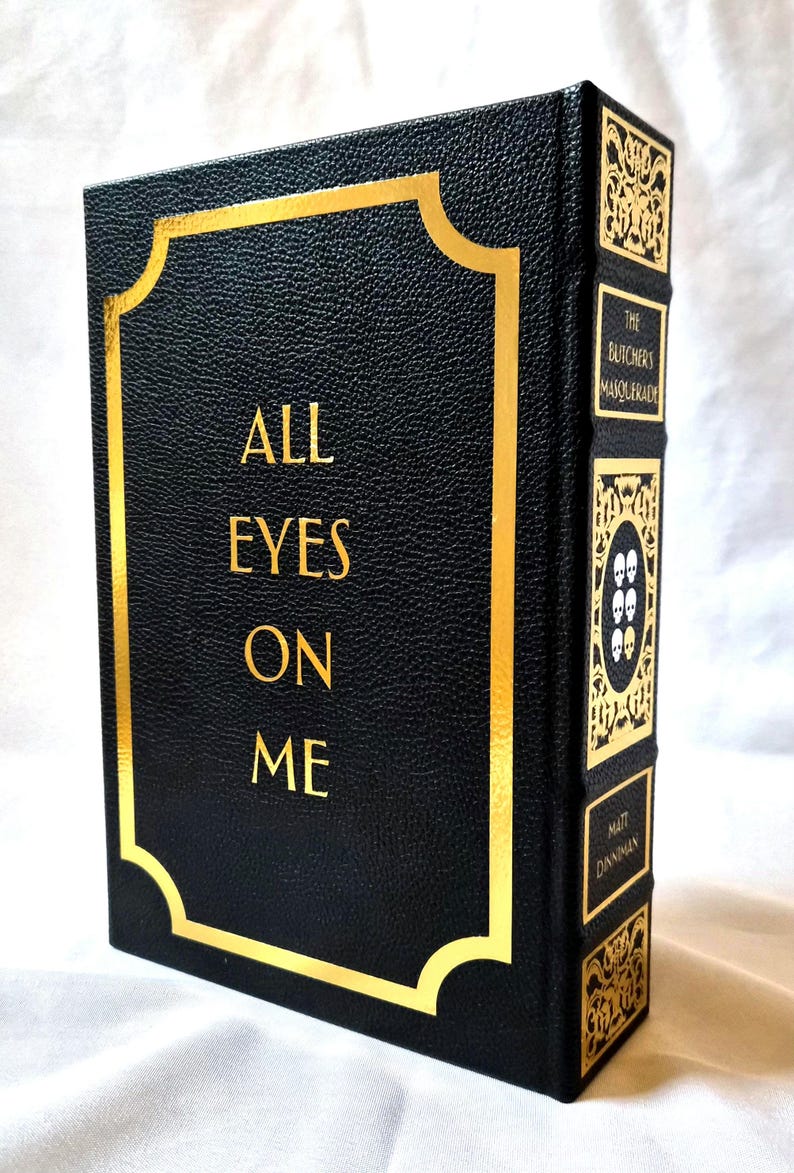 Puede incluir: Un libro de tapa dura negro con acabado texturizado y letras doradas en la portada que dicen ALL EYES ON ME. El lomo presenta paneles decorativos dorados, el t&iacute;tulo THE BUTCHERS MASQUERADE y el nombre del autor MATT DINNIMAN.