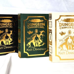 Puede incluir: Tres libros de tapa dura titulados Dungeon Crawler Carl de Matt Dinniman. Los libros presentan ilustraciones en l&aacute;mina dorada de una persona y un gato en las portadas, en colores negro, verde oscuro y crema con detalles en oro.