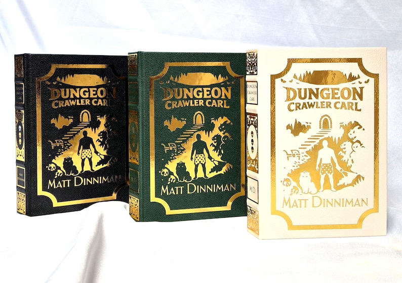 Puede incluir: Tres libros de tapa dura titulados Dungeon Crawler Carl de Matt Dinniman. Los libros presentan ilustraciones en l&aacute;mina dorada de una persona y un gato en las portadas, en colores negro, verde oscuro y crema con detalles en oro.