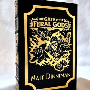 Puede incluir: Un libro de tapa dura negro titulado The Gate of the Feral Gods de Matt Dinniman. La portada presenta un estampado de l&aacute;mina dorada con una ilustraci&oacute;n de una persona con gafas protectoras haciendo paracaidismo junto a un gato.