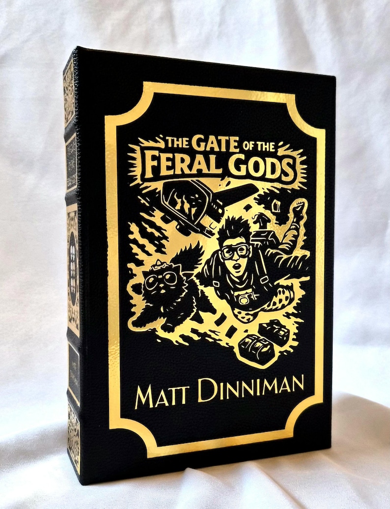 Puede incluir: Un libro de tapa dura negro titulado The Gate of the Feral Gods de Matt Dinniman. La portada presenta un estampado de l&aacute;mina dorada con una ilustraci&oacute;n de una persona con gafas protectoras haciendo paracaidismo junto a un gato.