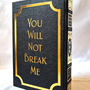 Puede incluir: Un libro de tapa dura negro con acabado texturizado y letras doradas que dicen "YOU WILL NOT BREAK ME" dentro de un borde dorado. El lomo presenta paneles decorativos dorados con iconos de calaveras y texto en orientaci&oacute;n vertical.