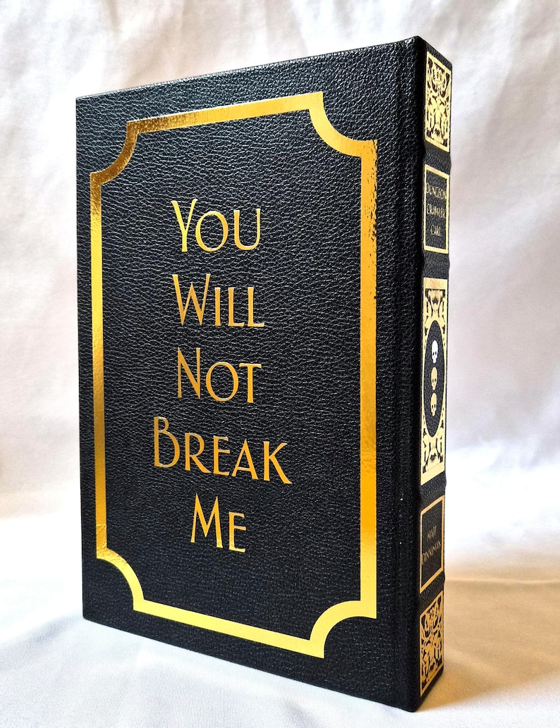Puede incluir: Un libro de tapa dura negro con acabado texturizado y letras doradas que dicen "YOU WILL NOT BREAK ME" dentro de un borde dorado. El lomo presenta paneles decorativos dorados con iconos de calaveras y texto en orientaci&oacute;n vertical.