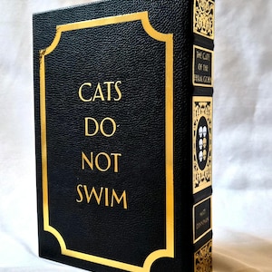 Puede incluir: Un libro de tapa dura negro con acabado texturizado y letras doradas que dicen CATS DO NOT SWIM en la portada. El lomo presenta filigrana dorada, cuatro iconos de calaveras y el texto THE CATE OF THE FERAL GOD y el nombre MATT DINNIMAN.