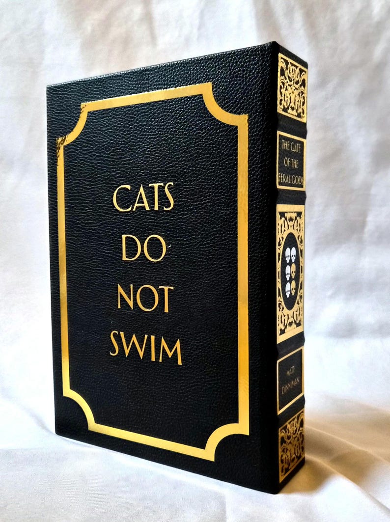 Puede incluir: Un libro de tapa dura negro con acabado texturizado y letras doradas que dicen CATS DO NOT SWIM en la portada. El lomo presenta filigrana dorada, cuatro iconos de calaveras y el texto THE CATE OF THE FERAL GOD y el nombre MATT DINNIMAN.
