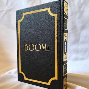 Puede incluir: Un libro de tapa dura negro con acabado texturizado y detalles en l&aacute;mina de oro. La portada presenta la palabra BOOM! en letras doradas dentro de un marco decorativo dorado. El lomo muestra patrones dorados ornamentados y tres iconos de calaveras.