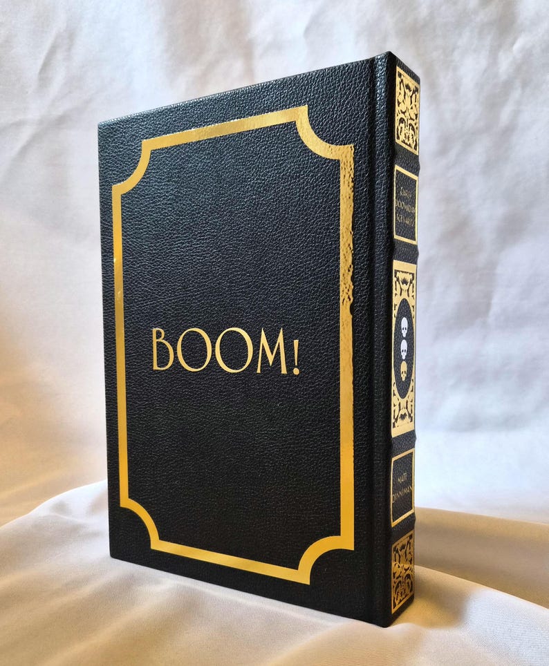 Puede incluir: Un libro de tapa dura negro con acabado texturizado y detalles en l&aacute;mina de oro. La portada presenta la palabra BOOM! en letras doradas dentro de un marco decorativo dorado. El lomo muestra patrones dorados ornamentados y tres iconos de calaveras.