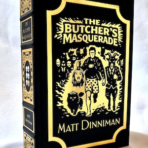Puede incluir: Un libro de tapa dura negro titulado The Butcher's Masquerade de Matt Dinniman con estampado de l&aacute;mina dorada. La portada presenta una ilustraci&oacute;n central de una persona sosteniendo una cabeza cortada, un gato y una figura con cabeza de cabra.