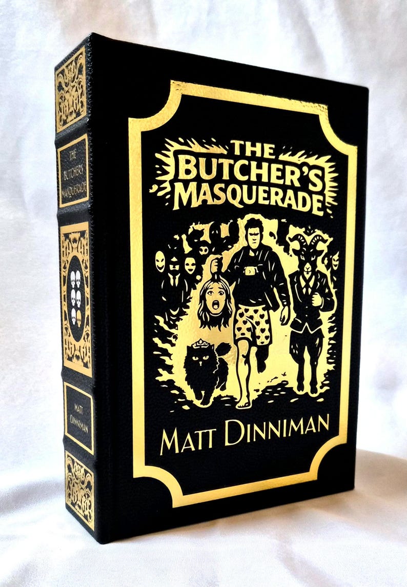 Puede incluir: Un libro de tapa dura negro titulado The Butcher's Masquerade de Matt Dinniman con estampado de l&aacute;mina dorada. La portada presenta una ilustraci&oacute;n central de una persona sosteniendo una cabeza cortada, un gato y una figura con cabeza de cabra.