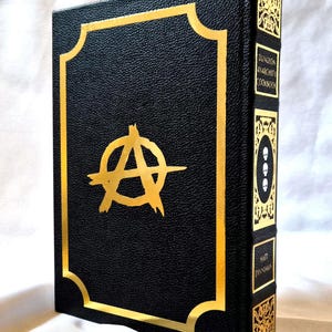 Puede incluir: Un libro de tapa dura negro titulado Dungeon Anarchist's Cookbook de Matt Dinniman. La portada presenta un s&iacute;mbolo de anarqu&iacute;a en l&aacute;mina dorada dentro de un borde dorado. El lomo muestra paneles decorativos dorados, tres calaveras y el autor.