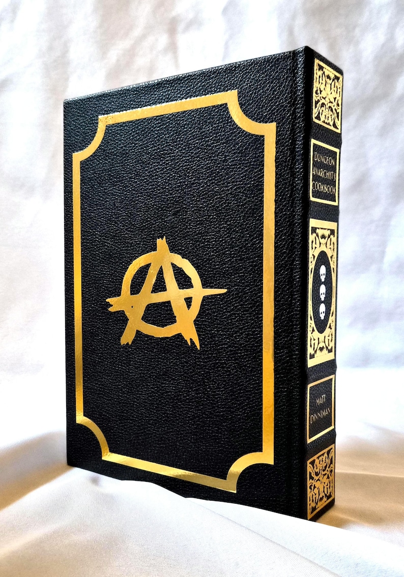 Puede incluir: Un libro de tapa dura negro titulado Dungeon Anarchist's Cookbook de Matt Dinniman. La portada presenta un s&iacute;mbolo de anarqu&iacute;a en l&aacute;mina dorada dentro de un borde dorado. El lomo muestra paneles decorativos dorados, tres calaveras y el autor.