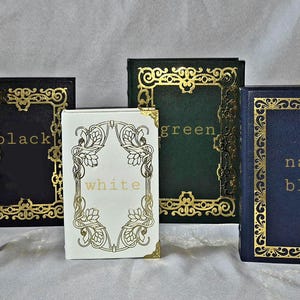 Puede incluir: Seis libros rectangulares dispuestos en fila, cada uno con un borde dorado ornamentado y el nombre del color impreso en letras doradas en el centro. Las portadas son marr&oacute;n, negro, blanco, verde, azul marino y rojo con filigranas.