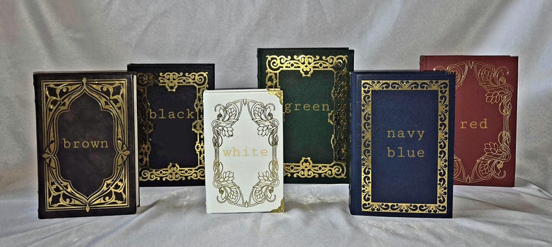 Puede incluir: Seis libros rectangulares dispuestos en fila, cada uno con un borde dorado ornamentado y el nombre del color impreso en letras doradas en el centro. Las portadas son marr&oacute;n, negro, blanco, verde, azul marino y rojo con filigranas.