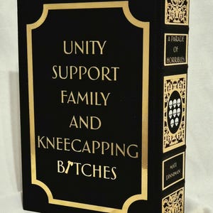 Puede incluir: Un libro negro con letras doradas en la portada que dice UNITY SUPPORT FAMILY AND KNEECAPPING BITCHES. El lomo presenta paneles decorativos dorados, el texto A PARADE OF HORRIBLES, un emblema de calavera y el nombre MATT DINNIMAN.