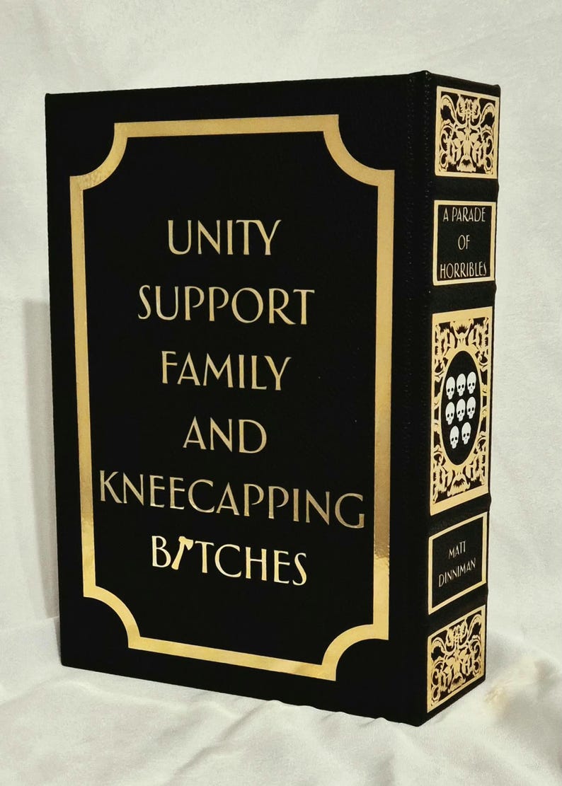Puede incluir: Un libro negro con letras doradas en la portada que dice UNITY SUPPORT FAMILY AND KNEECAPPING BITCHES. El lomo presenta paneles decorativos dorados, el texto A PARADE OF HORRIBLES, un emblema de calavera y el nombre MATT DINNIMAN.