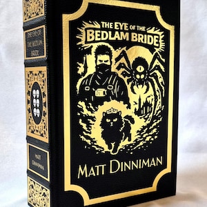 Puede incluir: Un libro de tapa dura negro titulado The Eye of the Bedlam Bride de Matt Dinniman. La portada presenta ilustraciones en l&aacute;mina dorada de una figura enmascarada, una ara&ntilde;a y un gato con corona. El lomo muestra el t&iacute;tulo y el autor en oro.