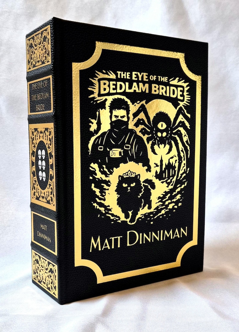 Puede incluir: Un libro de tapa dura negro titulado The Eye of the Bedlam Bride de Matt Dinniman. La portada presenta ilustraciones en l&aacute;mina dorada de una figura enmascarada, una ara&ntilde;a y un gato con corona. El lomo muestra el t&iacute;tulo y el autor en oro.