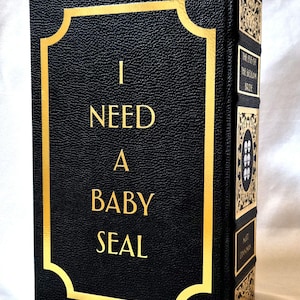 Puede incluir: Un libro negro con cubierta de textura similar al cuero que presenta letras doradas con el texto I NEED A BABY SEAL dentro de un borde dorado. El lomo muestra paneles decorativos dorados, el t&iacute;tulo THE EYE OF THE BEDLAM BRIDE y el autor.