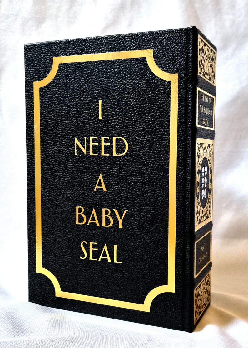 Puede incluir: Un libro negro con cubierta de textura similar al cuero que presenta letras doradas con el texto I NEED A BABY SEAL dentro de un borde dorado. El lomo muestra paneles decorativos dorados, el t&iacute;tulo THE EYE OF THE BEDLAM BRIDE y el autor.